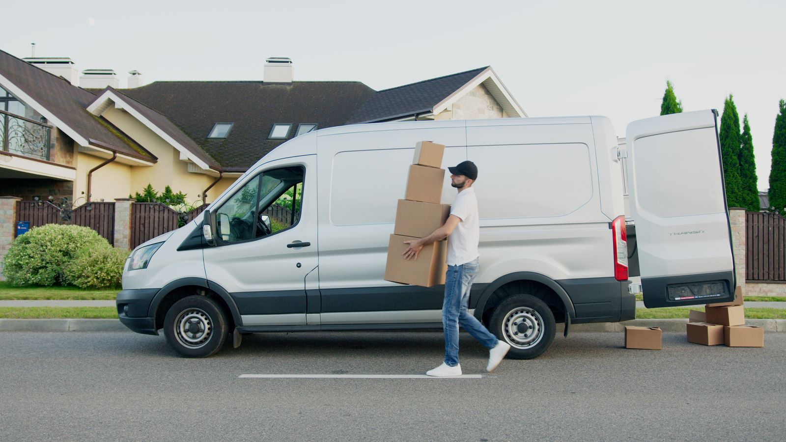 Courier carrying stacked cardboard boxes from a white Transit van outside a house